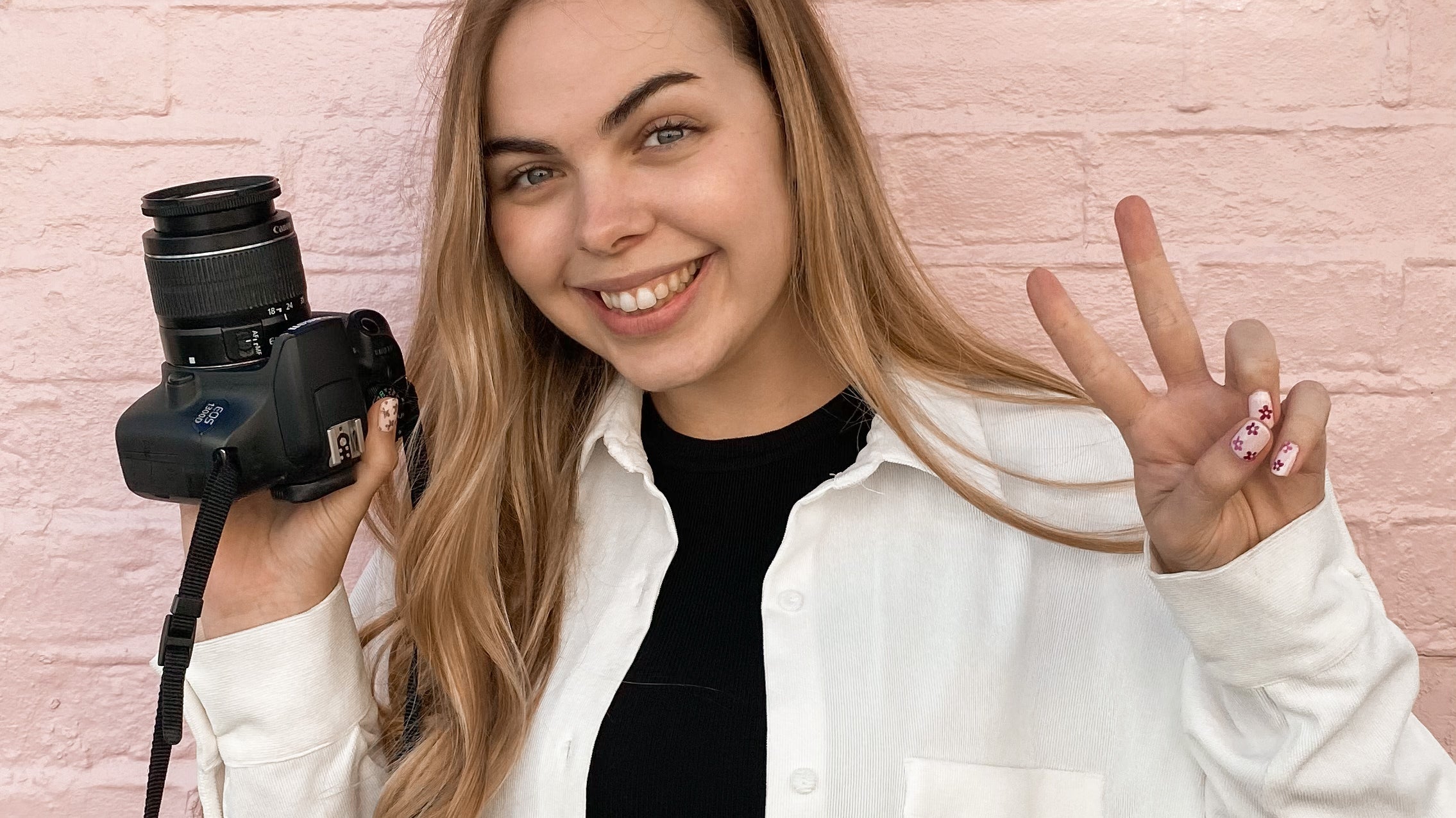 Meet the team - Elsa - Elsa standing in front of a pink wall holding her camera in one hand a doing a peace sign with the other.