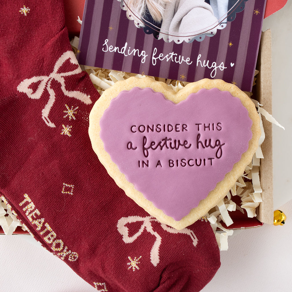 A heart-shaped sugar cookie with lavender icing and the text 'CONSIDER THIS a festive hug IN A BISCUIT' sits in a gift box with shredded paper. A burgundy knit sock with white bows and gold snowflake motifs is partly visible, along with a purple striped card reading 'Sending festive hugs'.