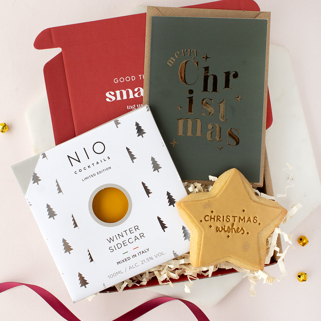 A curated gift set featuring a NIO Winter Sidecar cocktail mix box, a dark green Christmas greeting card with gold foil lettering, a red box insert, and a star-shaped Christmas cookie on a white background.