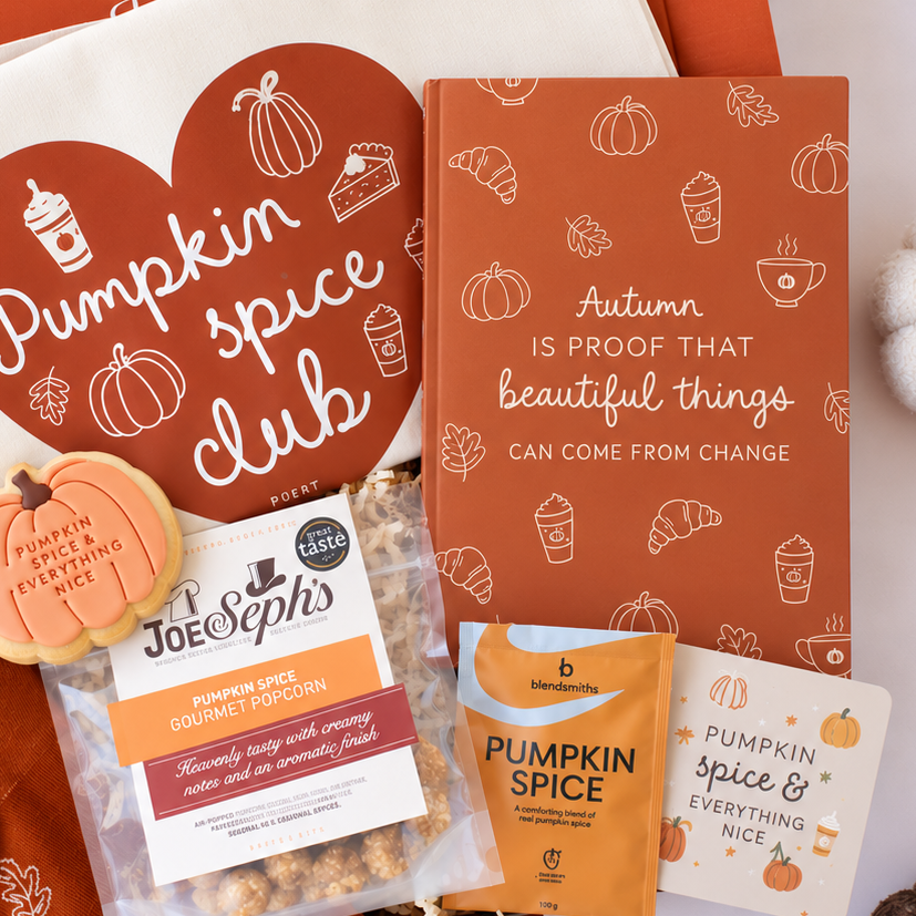 A styled autumn gift set featuring a white bag with a large red heart that reads 'Pumpkin spice club,' an orange book with white line drawings, a cookie labeled 'Pumpkin Spice & Everything Nice,' a bag of Joe's Pops pumpkin spice gourmet popcorn, a Blendworks pumpkin spice snack pouch, and a card reading 'Pumpkin Spice & Everything Nice.'.