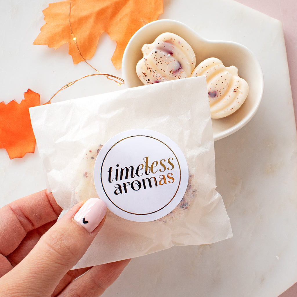 Hand holding a small bag of wax melts with a bowl of two pumpkin shaped wax melts