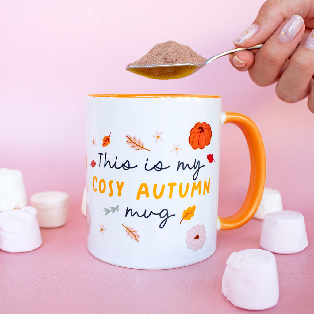 A mug with the words This is my cosy Autumn mug with a hand holding a spoonful of drinking chocolate powder