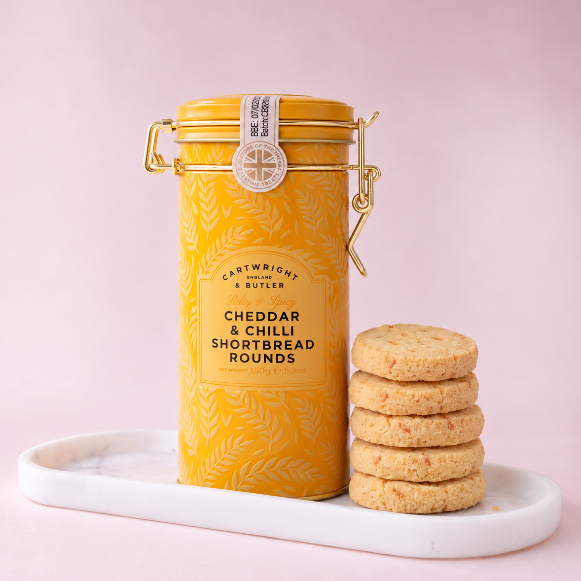 A yellow cylindrical metal tin of Castwright & Butler Cheddar & Chilli Shortbread Rounds stands on a white marble tray next to a stack of four golden-brown shortbread cookies, set against a solid pink background.