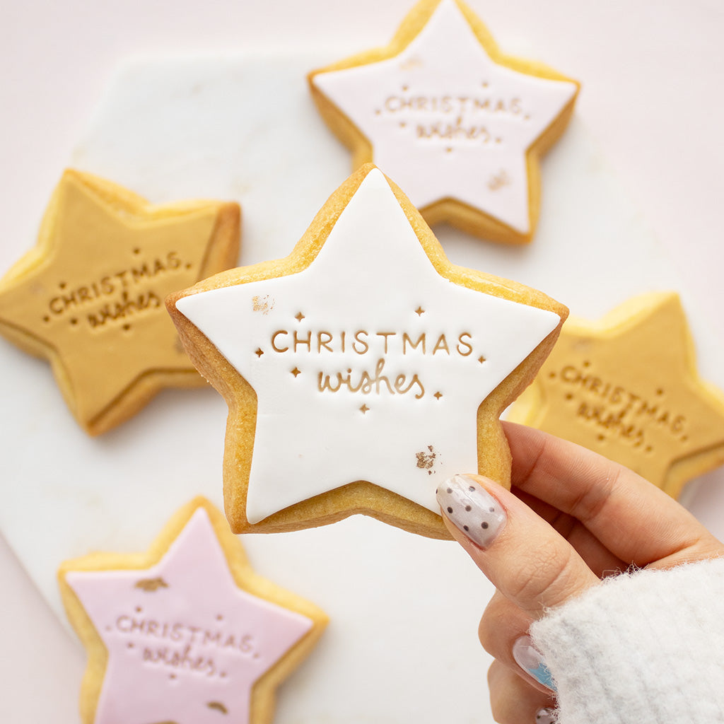 A hand with white and silver nail polish holds a star-shaped sugar cookie decorated with white icing and gold lettering that reads 'Christmas wishes'. In the background, other star-shaped cookies in gold, pink, and white icing are arranged on a white marble surface.