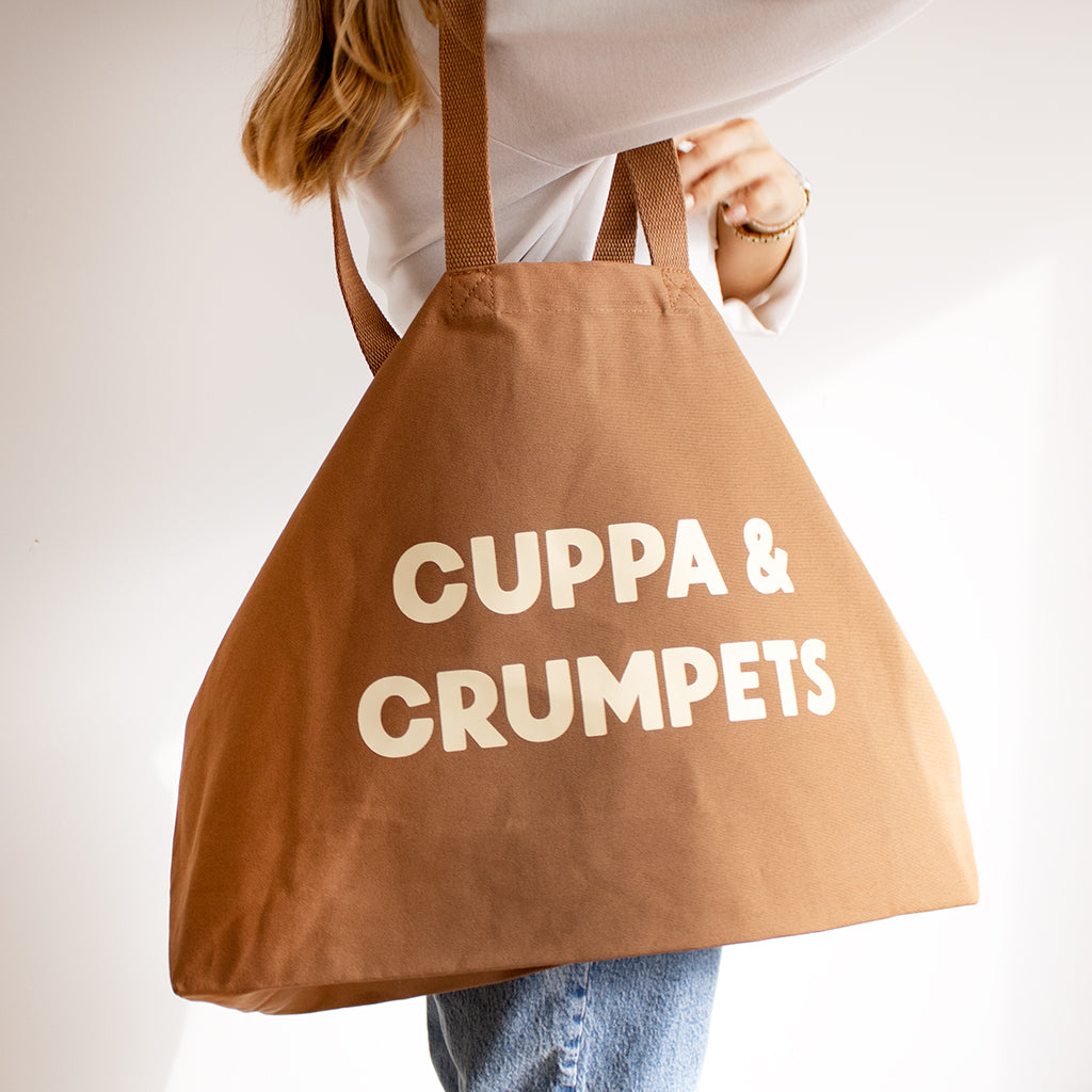 A person holding a large brown canvas tote bag with the words 'CUPPA & CRUMPETS' printed in bold cream letters, shown against a plain light background.