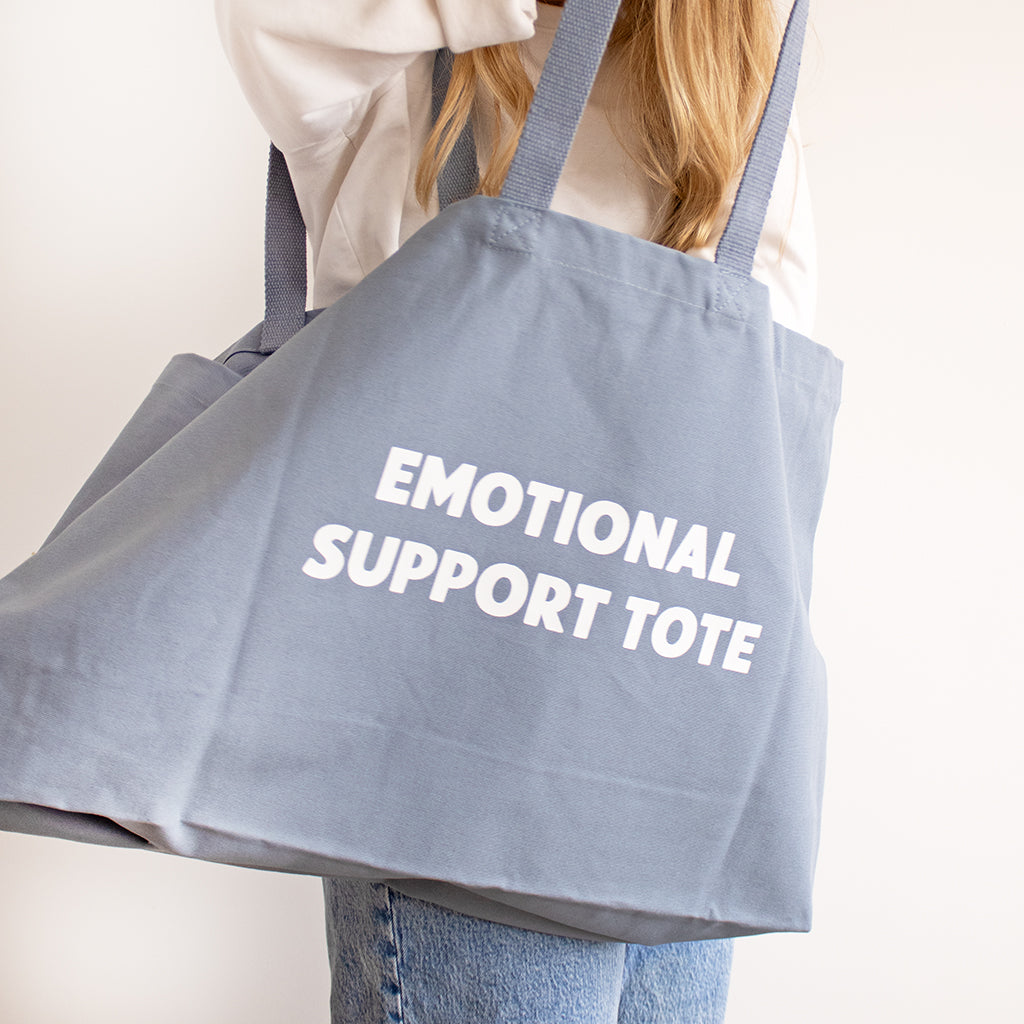 A person carrying a large dusty blue canvas tote bag with the words 'EMOTIONAL SUPPORT TOTE' printed in white capital letters on the front, set against a plain off-white background.
