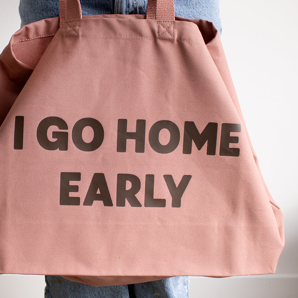 A large dusty pink rose canvas tote bag with the words 'I GO HOME EARLY' printed in bold brown letters.