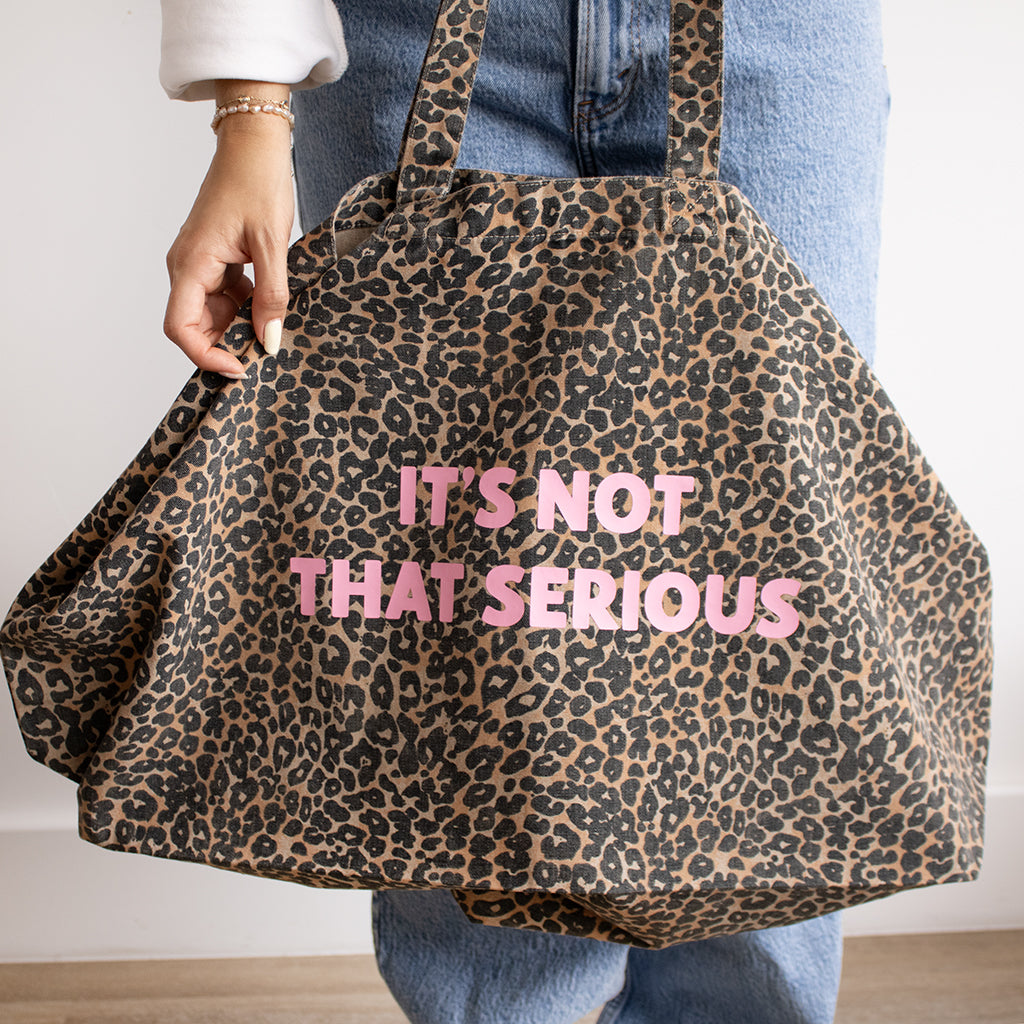 A person wearing blue jeans and a white top holds up a large leopard print tote bag with pink text that reads 'IT'S NOT THAT SERIOUS'.