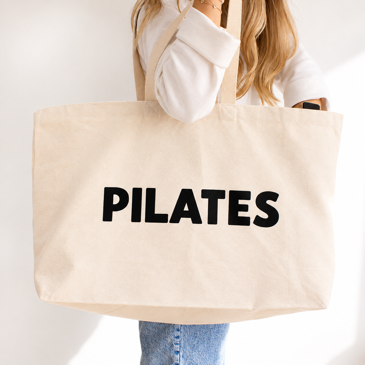A person holding a large natural beige canvas tote bag with the word 'PILATES' printed in bold black letters across the front, against a clean white background.