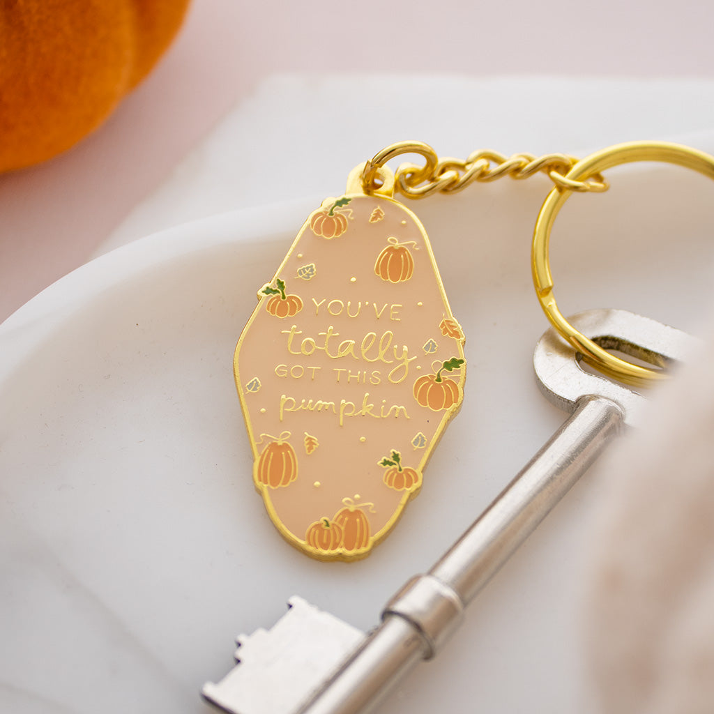 A gold-tone metal keychain with a peach-colored oval charm featuring pumpkin illustrations and the phrase 'YOU'VE totally GOT THIS pumpkin' in gold lettering, shown with a key on a white marble surface.