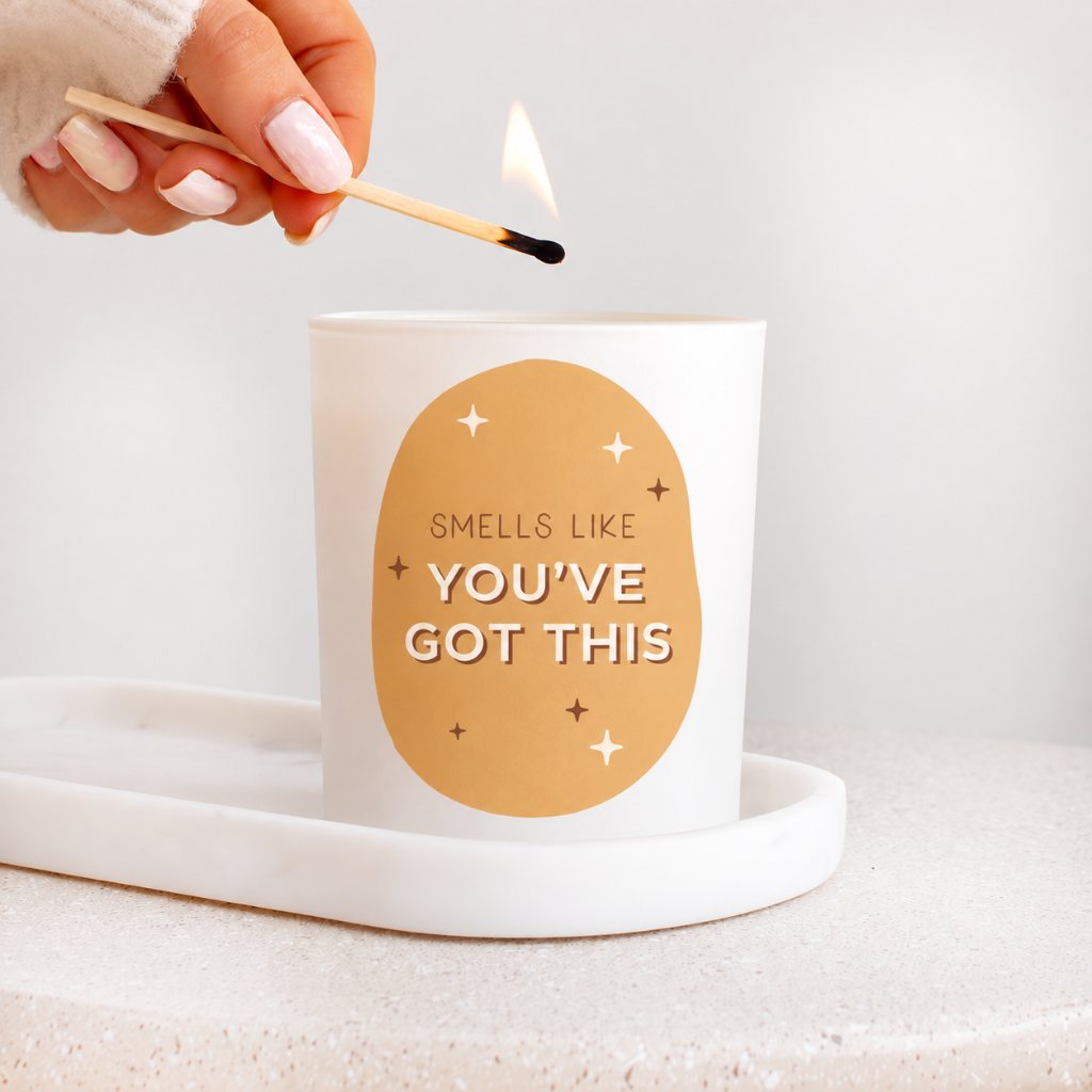 A white candle in a white glass holder sits on a white tray, with a hand lighting a match above it. The front of the candle features a tan oval graphic with the text 'SMELLS LIKE YOU'VE GOT THIS' and small star accents.