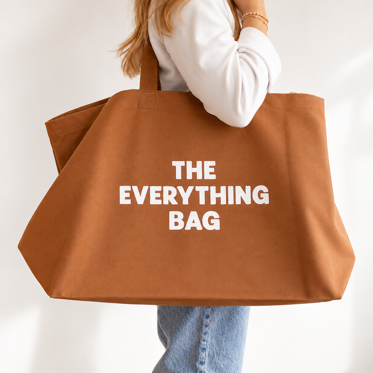 A person holding a large brown canvas tote bag with the words 'THE EVERYTHING BAG' printed in bold white letters on the front, set against a clean white background.