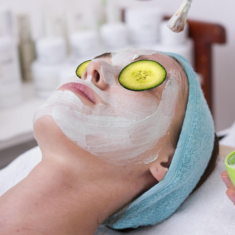 Person receiving a facial treatment with cucumbers on eyes and a bowl of cream.