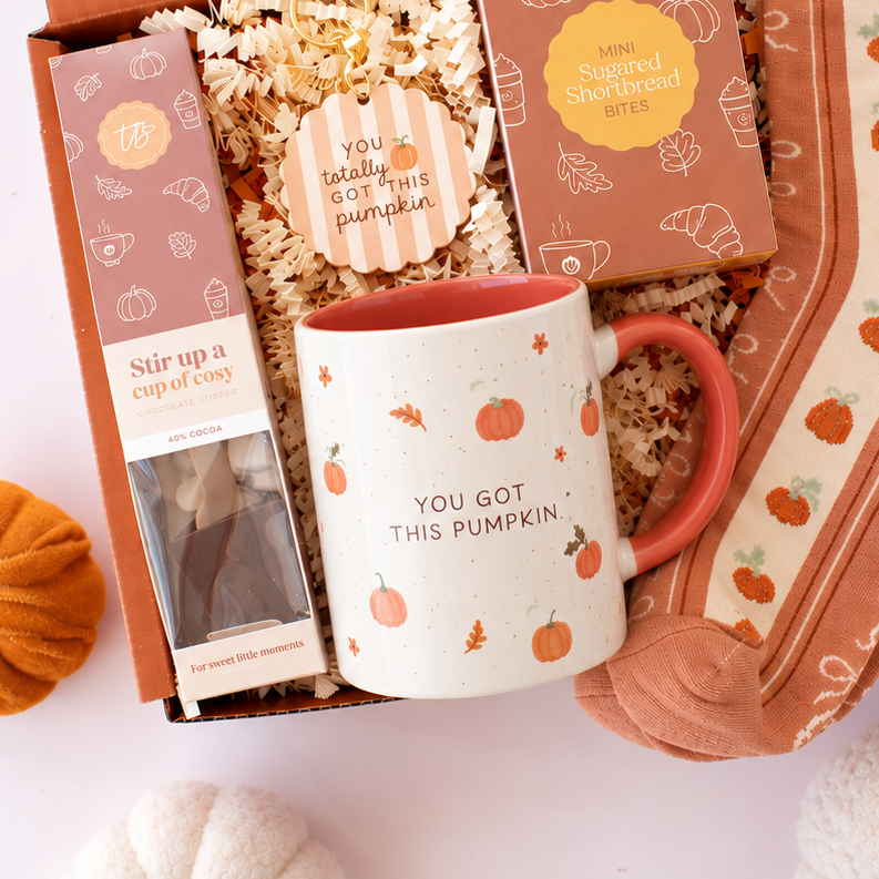 An autumn-themed gift box set from Treatbox, featuring a white ceramic mug with a pumpkin design, patterned socks, a pumpkin-shaped candle, and a tea bag, arranged in an open orange box with decorative pumpkins on a white background.