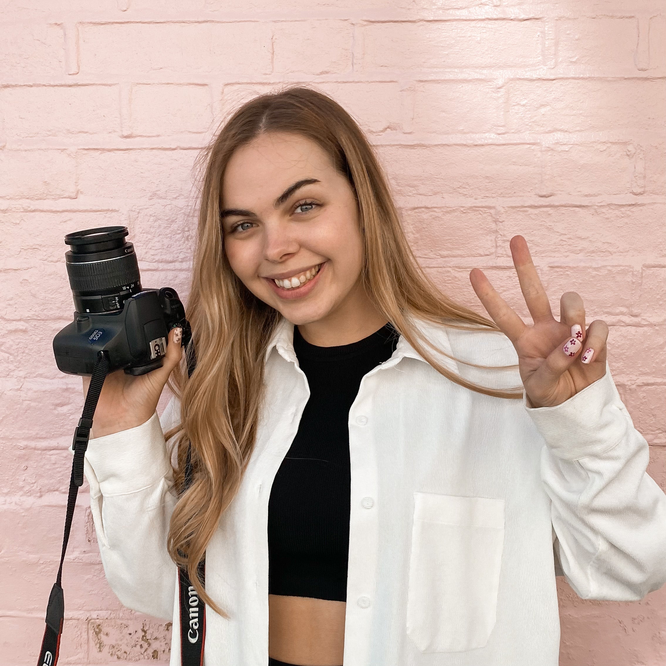Meet the team - Elsa - Elsa standing in front of a pink wall holding her camera in one hand a doing a peace sign with the other.