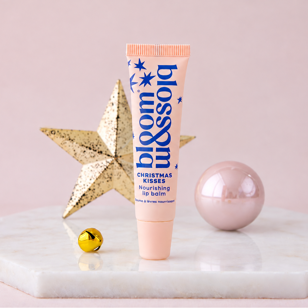 A squeeze tube of Blossom Kisses Christmas Kisses nourishing lip balm stands upright on a white marble slab. The tube is peach-pink with blue text and star graphics.