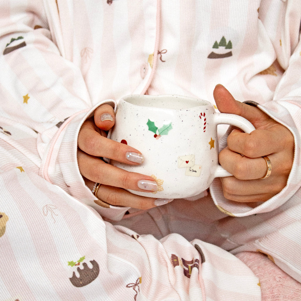 A person wearing light pink and white striped pajamas with Christmas-themed prints, holding a white ceramic mug with holiday motifs.