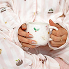 A person wearing light pink and white striped pajamas with Christmas-themed prints, holding a white ceramic mug with holiday motifs.