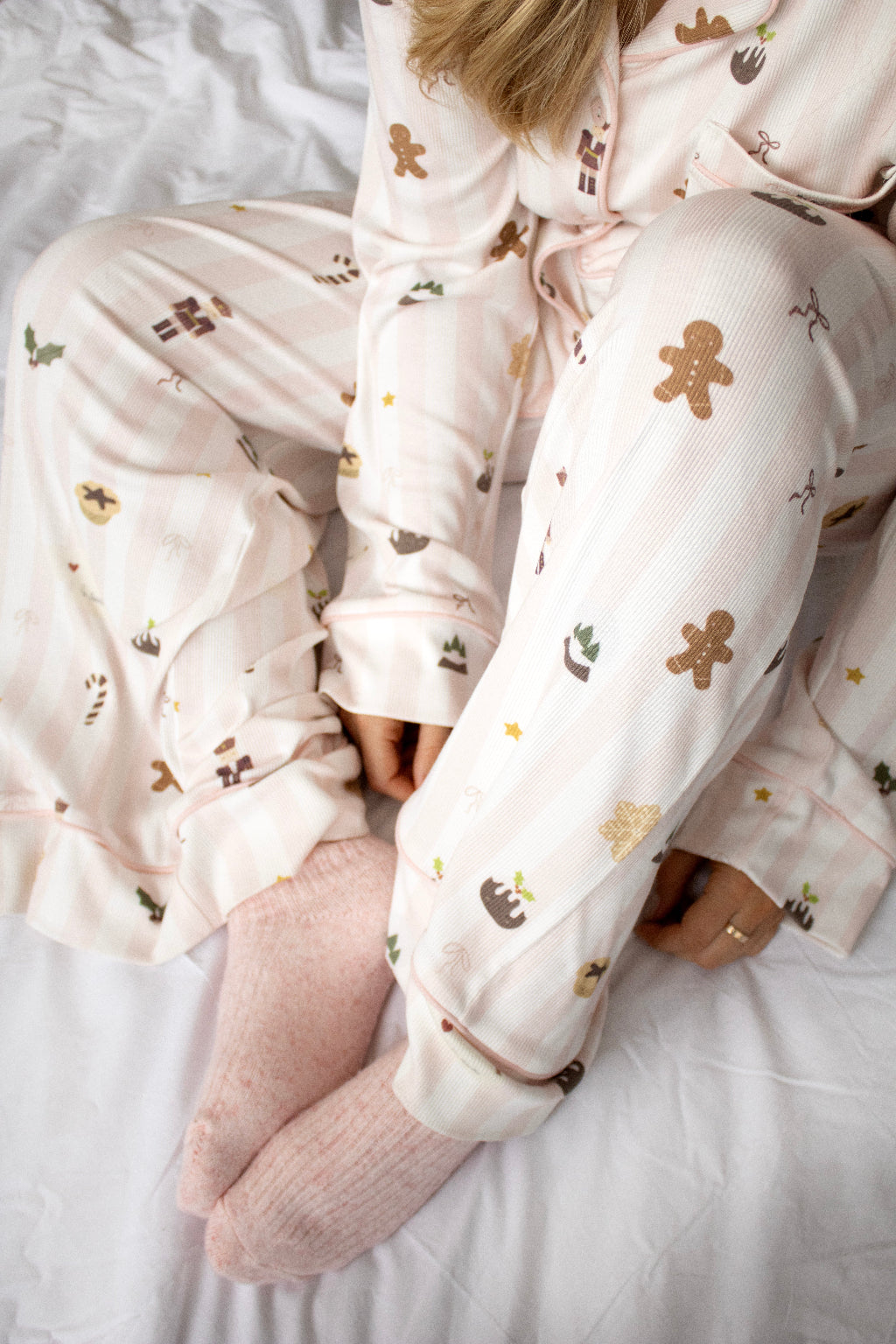 A person wearing light pink pajama pants and matching top with a gingerbread-themed print, sitting on white bedding with pink socks.