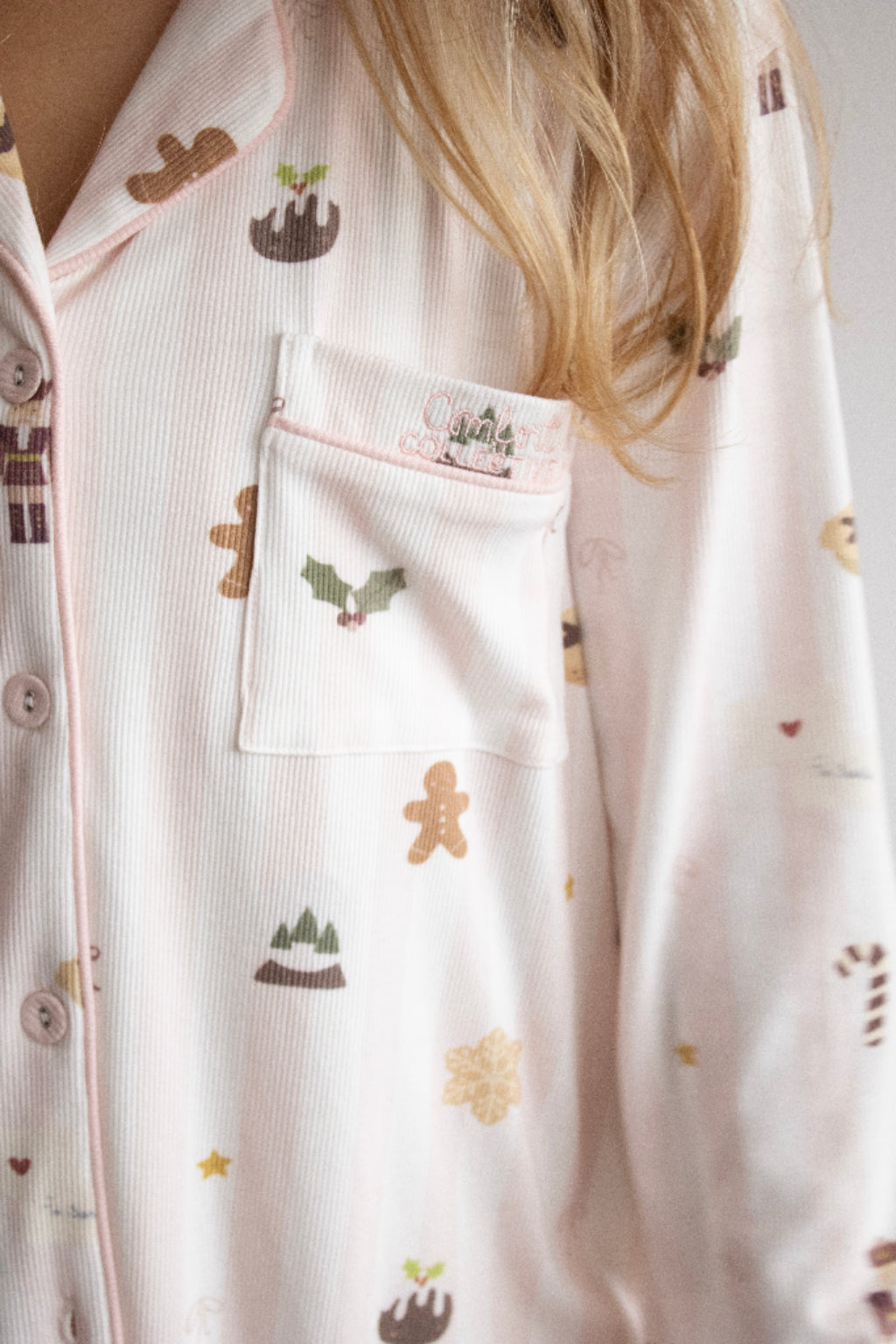 Close-up of a person wearing light pink ribbed pajama top with a holiday print, featuring gingerbread men, holly, stars, and a chest pocket with a matching patch.