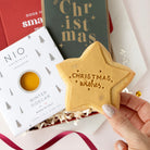 A hand holds a star-shaped gingerbread cookie with 'CHRISTMAS wishes' written on it, set against a festive gift box background with NIO Cocktails Winter Sidcar packaging and Christmas decorations.