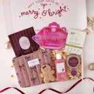 A curated gift box set with multiple beauty and self-care items arranged in a white box with shredded paper. Visible items include a pink quilted pouch with black bow print, a pink lip mask packet, a gingerbread cookie-shaped item, a pink bottle, a round tin, a striped packet, and a cream facial mask packet.