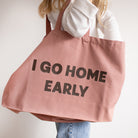 A person wearing a white top and blue jeans carries a large dusty pink rose canvas tote bag with the words 'I GO HOME EARLY' printed in bold brown letters.