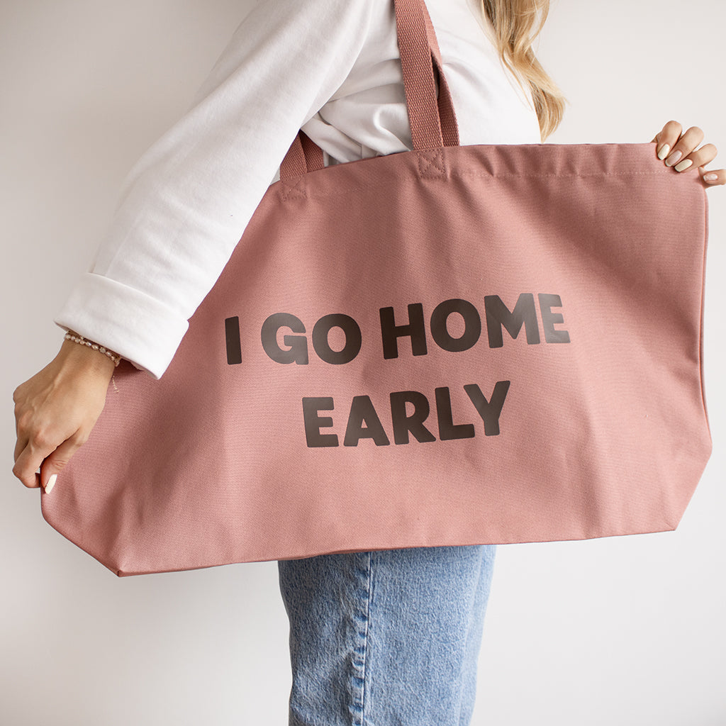 A person wearing a white top and blue jeans carries a large dusty pink rose canvas tote bag with the words 'I GO HOME EARLY' printed in bold brown letters.