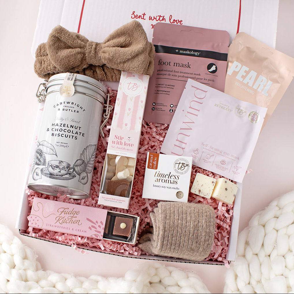 A curated gift box filled with assorted self-care and food items, photographed from above on a soft pink background. The box contains a pink bow, a tin of hazelnut and chocolate biscuits, a foot mask packet, a soap box, a pouch labeled PEARL, a small box of timeless aromas, a chocolate, and a folded knit item, all arranged in pink shredded paper.