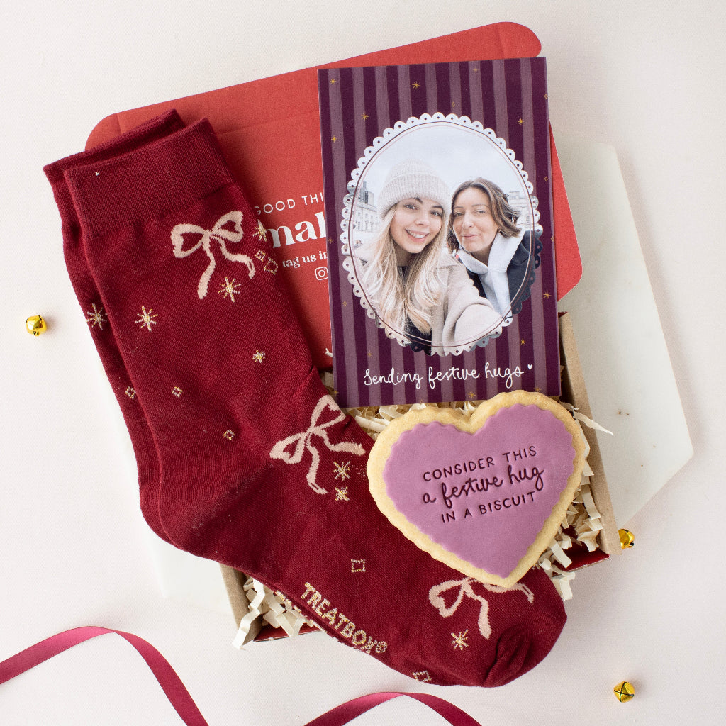 A holiday gift set featuring a pair of deep red crew socks with white bow and star patterns, a custom photo card, a heart-shaped sugar cookie, and a red envelope, arranged in a box with white tissue paper.