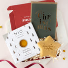 A curated gift set featuring a NIO Winter Sidecar cocktail mix box, a dark green Christmas greeting card with gold foil lettering, a red box insert, and a star-shaped Christmas cookie on a white background.