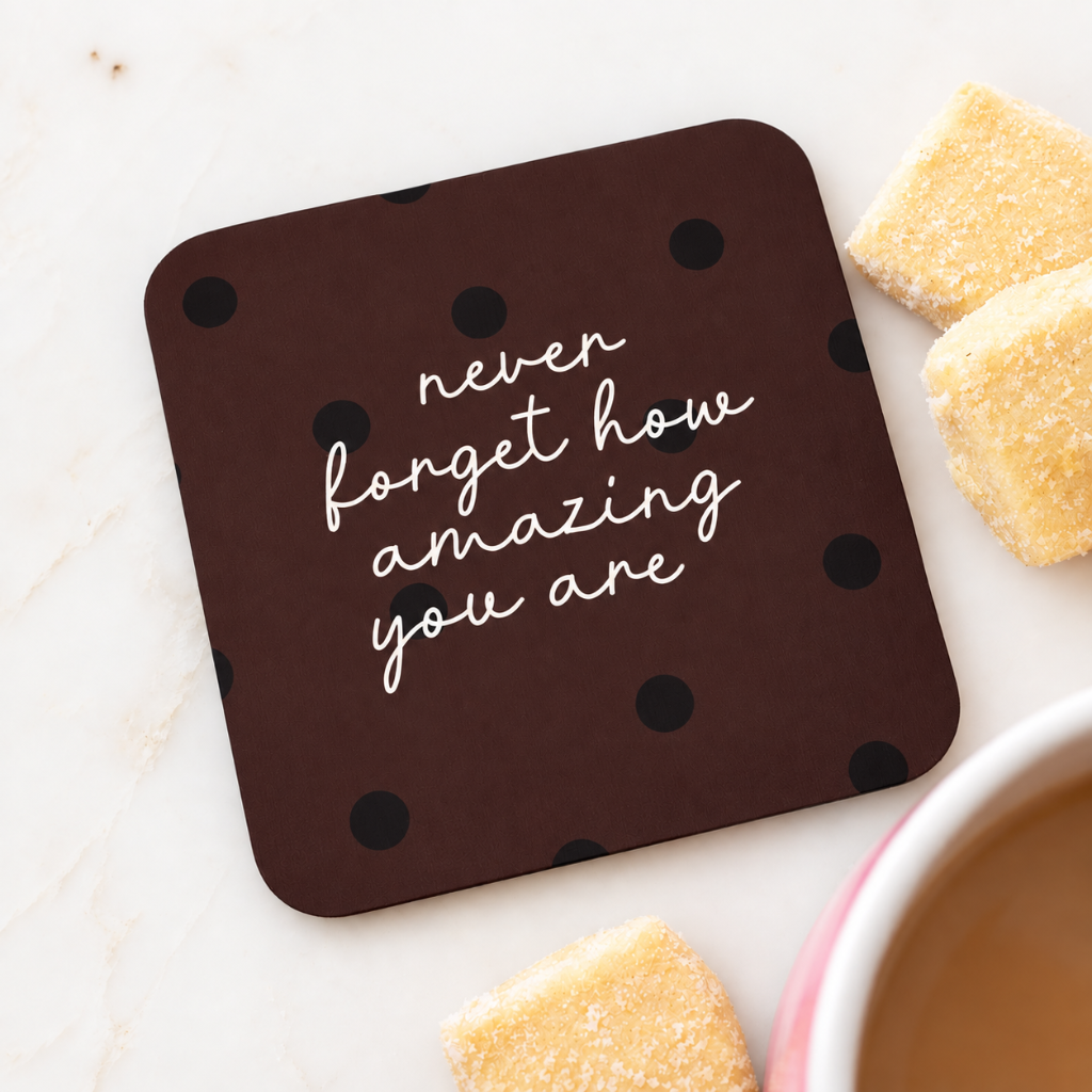 A square dark brown coaster with black polka dots and the phrase 'never forget how amazing you are' in white script, styled on a light marble surface with sugar cubes and a cup of coffee nearby.