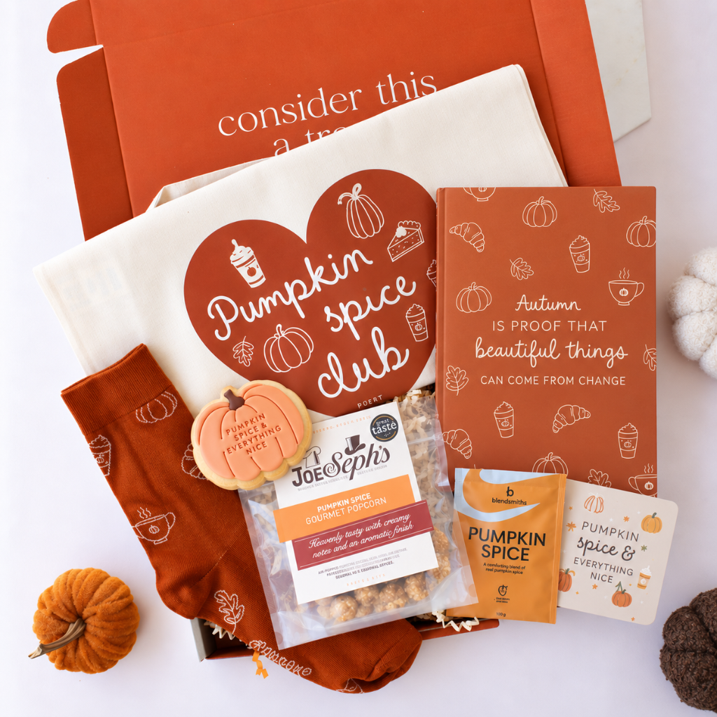 A styled flat-lay of a fall-themed gift box set on a white background, featuring an orange box, a cream tumbler with a brown heart graphic reading 'Pumpkin spice club,' rust-colored socks, a small pumpkin, a packet of Joe's Spics pumpkin spice coffee, and a card that says 'Autumn is proof that beautiful things can come from change.'.