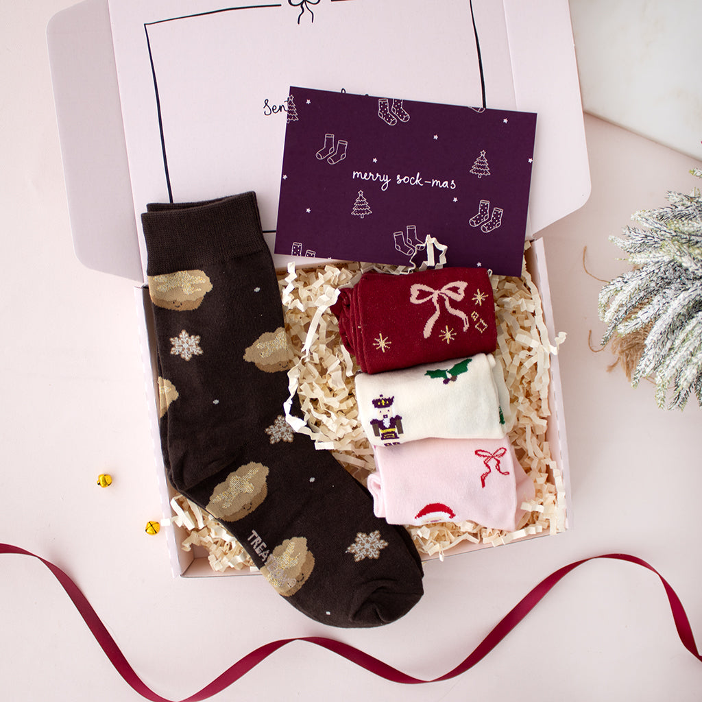A Christmas-themed sock gift set in an open white box filled with shredded paper. One dark brown sock with gold and white holiday motifs is laid flat, while three smaller socks in red, cream, and light pink are stacked inside.