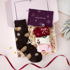 A Christmas-themed sock gift set in an open white box filled with shredded paper. One dark brown sock with gold and white holiday motifs is laid flat, while three smaller socks in red, cream, and light pink are stacked inside.