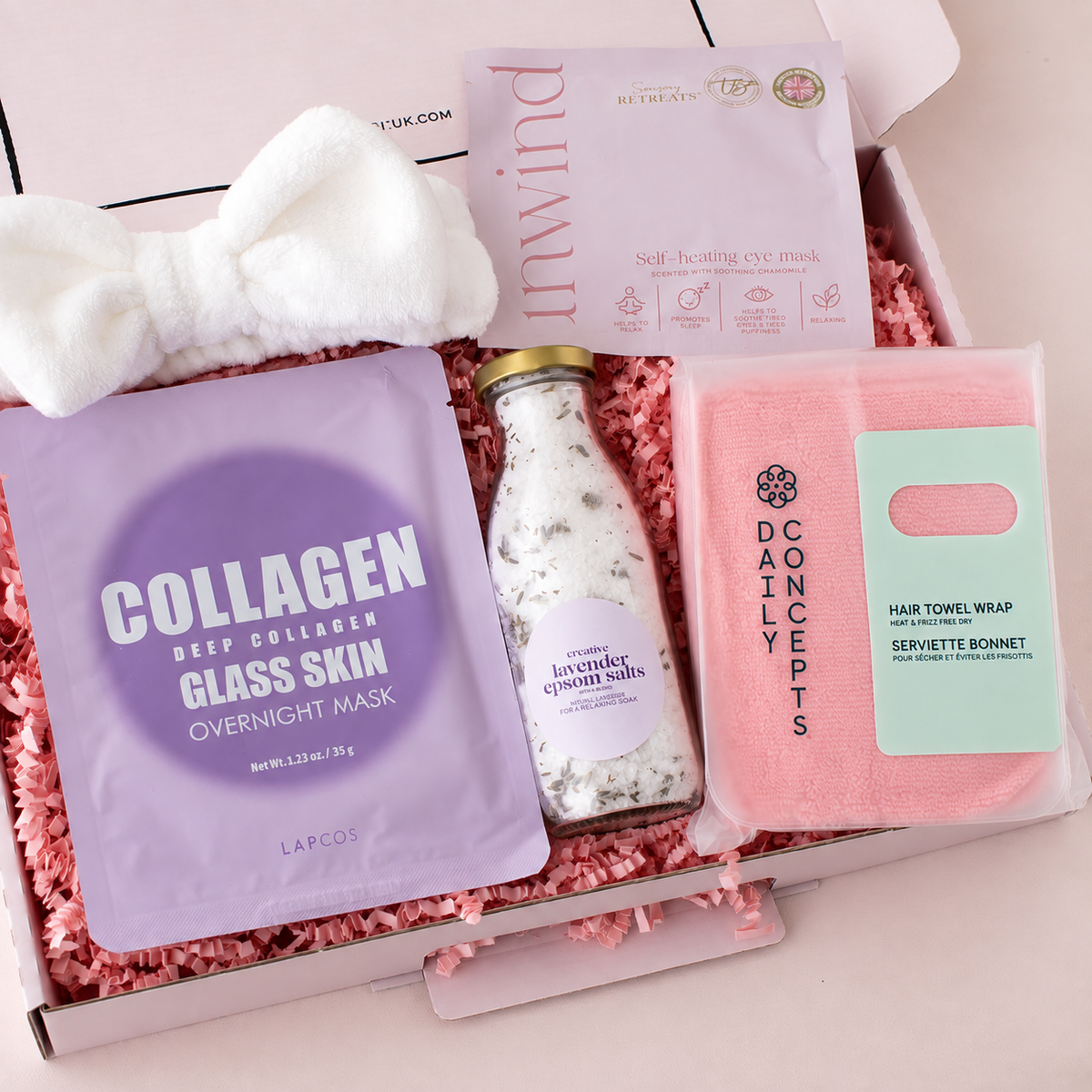 A curated skincare gift box filled with pink shredded paper, containing a white bow headband, a lavender Laapos Collagen Deep Collagen Glass Skin Overnight Mask packet, a bottle of lavender Epsom bath salts, a pink Concepts by Dain Concept Hair Towel Wrap, and an Inwind Self-Healing eye mask.