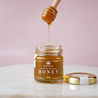 A small glass jar of Maters & co Infused Natural Honey sits on a light marble surface with honey dripping from a wooden dipper above it. The gold lid rests beside the jar against a soft pink background.