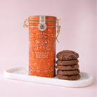 An orange metal canister of Cartwright Butler Chocolate Birthday Biscuits sits on a white marble tray next to a stack of chocolate chip cookies against a soft pink background.