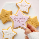 A hand with white and silver nail polish holds a star-shaped sugar cookie decorated with white icing and gold lettering that reads 'Christmas wishes'. In the background, other star-shaped cookies in gold, pink, and white icing are arranged on a white marble surface.