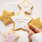 A hand with white and silver nail polish holds a star-shaped sugar cookie decorated with white icing and gold lettering that reads 'Christmas wishes'. In the background, other star-shaped cookies in gold, pink, and white icing are arranged on a white marble surface.