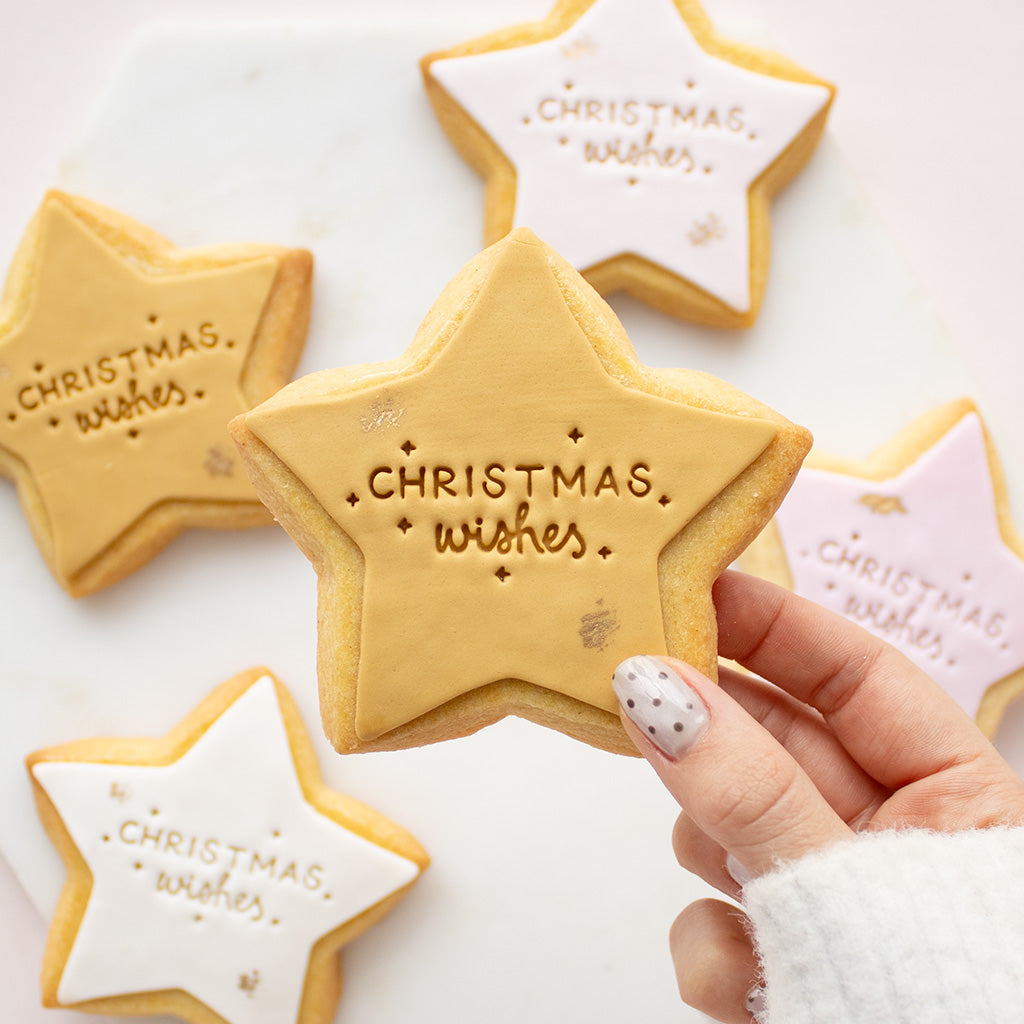 A hand with white and silver nail polish holds a star-shaped sugar cookie decorated with white icing and gold lettering that reads 'Christmas wishes'. In the background, other star-shaped cookies in gold, pink, and white icing are arranged on a white marble surface.