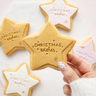 A hand with white and silver nail polish holds a star-shaped sugar cookie decorated with white icing and gold lettering that reads 'Christmas wishes'. In the background, other star-shaped cookies in gold, pink, and white icing are arranged on a white marble surface.