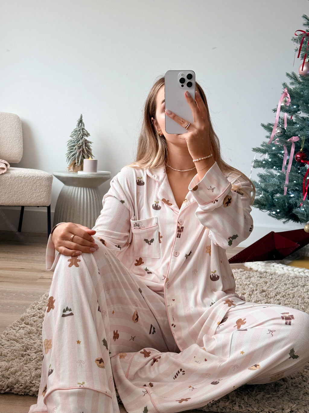A woman sitting on a shag rug takes a mirror selfie wearing a light pink pajama set with a holiday print of gingerbread cookies, candy canes, and snowflakes. The background shows a cozy living room with a Christmas tree and a small side table.