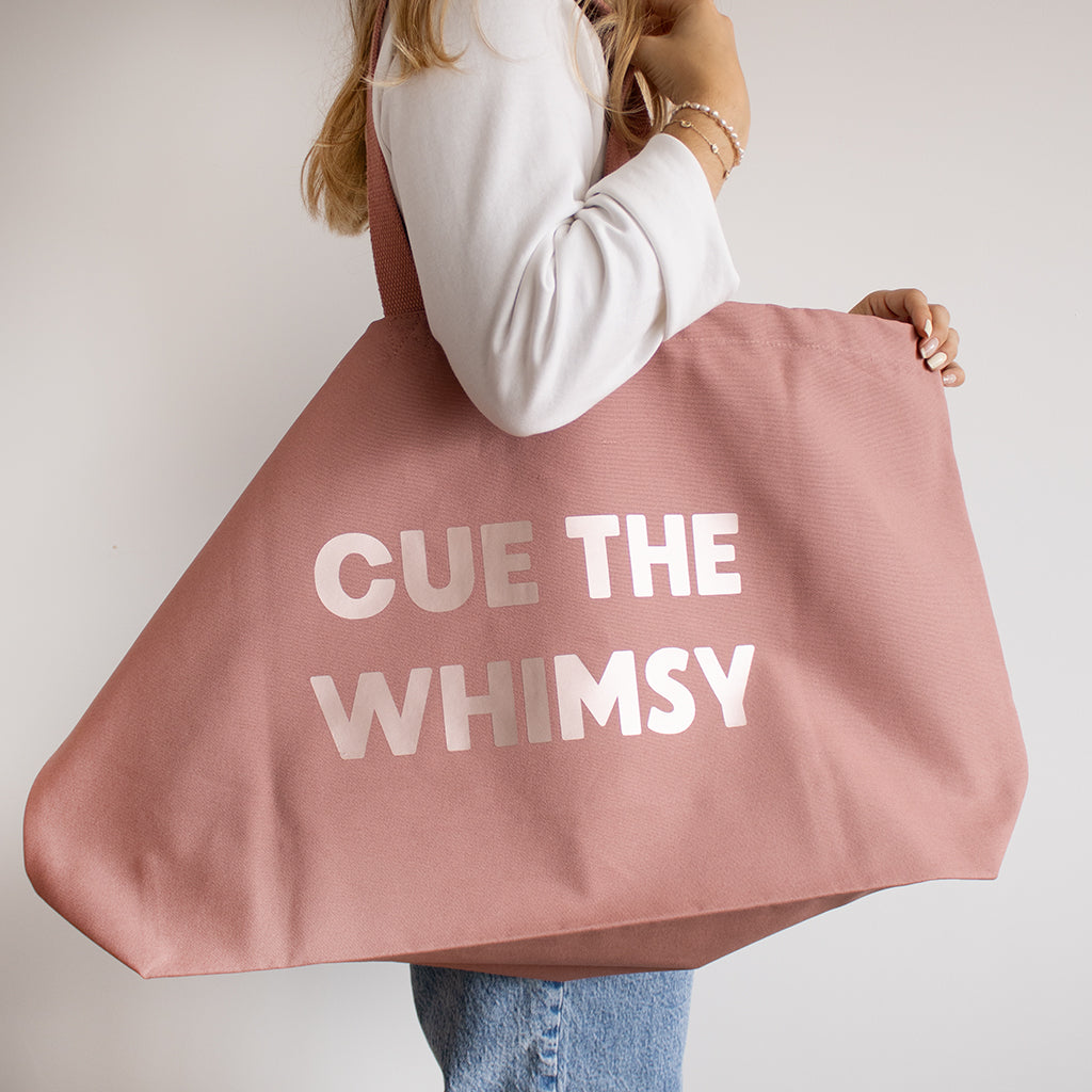 A person holding a large dusty pink canvas tote bag with the text 'CUE THE WHIMSY' printed in rose gold.