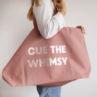 A person holding a large dusty pink canvas tote bag with the text 'CUE THE WHIMSY' printed in rose gold.