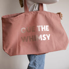 A person holding a large dusty pink canvas tote bag with the text 'CUE THE WHIMSY' printed in rose gold.