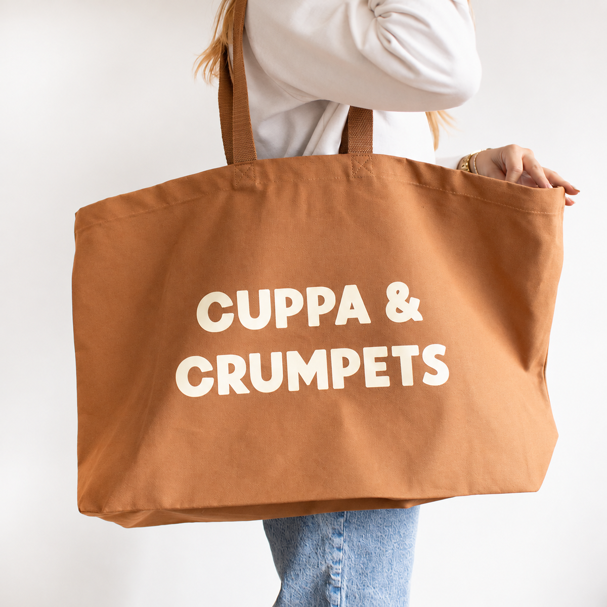 A person holding a large brown canvas tote bag with the words 'CUPPA & CRUMPETS' printed in bold cream letters, shown against a plain light background.