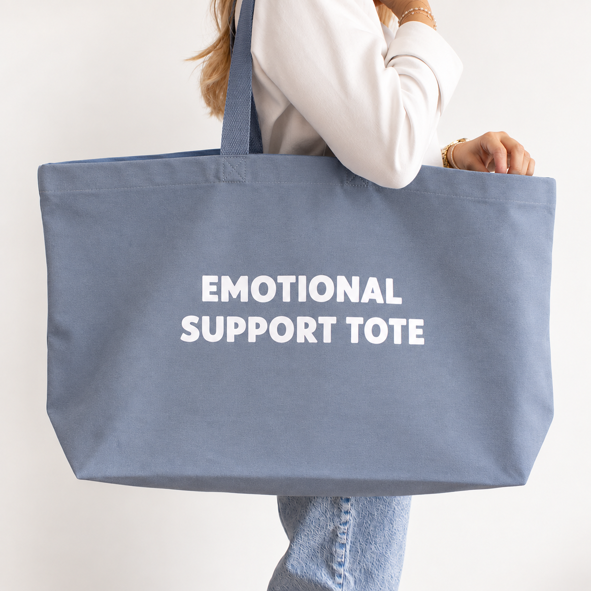 A person carrying a large dusty blue canvas tote bag with the words 'EMOTIONAL SUPPORT TOTE' printed in white capital letters on the front, set against a plain off-white background.
