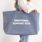 A person carrying a large dusty blue canvas tote bag with the words 'EMOTIONAL SUPPORT TOTE' printed in white capital letters on the front, set against a plain off-white background.