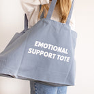 A person carrying a large dusty blue canvas tote bag with the words 'EMOTIONAL SUPPORT TOTE' printed in white capital letters on the front, set against a plain off-white background.